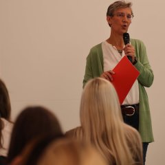 Eine Frau steht mit einem Mikro in einem Raum. Im Vordergrund sieht man Köpfe.