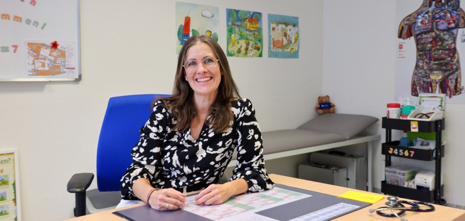 Dr. Stephanie Göpfert, Ärztin des Kinder- und Jugendärztlichen Dienstes, sitzt lächelnd an ihrem Schreibtisch im Gesundheitsamt Hersfeld-Rotenburg. Im Hintergrund sind kindgerechte Poster und medizinisches Equipment zu sehen.
