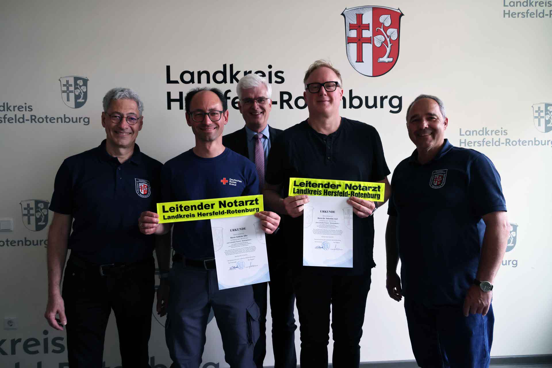 Fünf Männer stehen nebeneinander vor einer Wand mit dem Logo des Landkreises Hersfeld-Rotenburg. Zwei der Männer in der Mitte halten jeweils eine Urkunde und ein gelbes Schild mit der Aufschrift „Leitender Notarzt Landkreis Hersfeld-Rotenburg“ in der Hand.