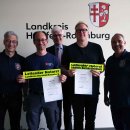 Fünf Männer stehen nebeneinander vor einer Wand mit dem Logo des Landkreises Hersfeld-Rotenburg. Zwei der Männer in der Mitte halten jeweils eine Urkunde und ein gelbes Schild mit der Aufschrift „Leitender Notarzt Landkreis Hersfeld-Rotenburg“ in der Hand.