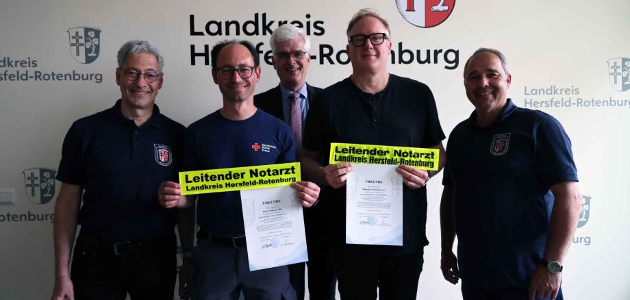 Fünf Männer stehen nebeneinander vor einer Wand mit dem Logo des Landkreises Hersfeld-Rotenburg. Zwei der Männer in der Mitte halten jeweils eine Urkunde und ein gelbes Schild mit der Aufschrift „Leitender Notarzt Landkreis Hersfeld-Rotenburg“ in der Hand.