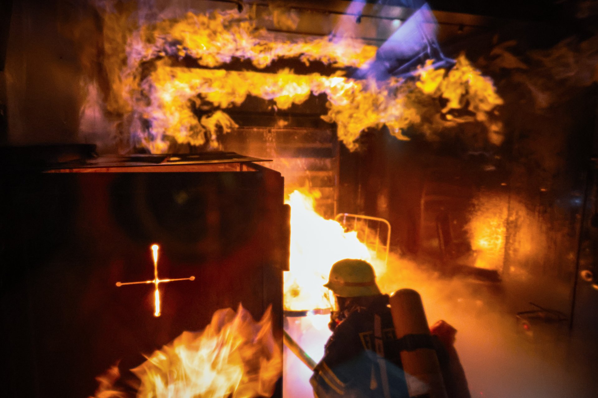 Ein Feuerwehrmann in voller Schutzausrüstung mit Atemschutzgerät befindet sich in einem brennenden Raum. Über ihm schlagen helle Flammen an der Decke entlang, dichter Rauch steigt auf. Die Szene zeigt eine realistische Trainingssituation zur Brandbekämpfung in einer mobilen Brandsimulationsanlage.