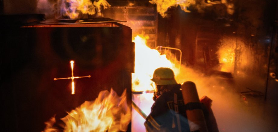 Ein Feuerwehrmann in voller Schutzausrüstung mit Atemschutzgerät befindet sich in einem brennenden Raum. Über ihm schlagen helle Flammen an der Decke entlang, dichter Rauch steigt auf. Die Szene zeigt eine realistische Trainingssituation zur Brandbekämpfung in einer mobilen Brandsimulationsanlage.