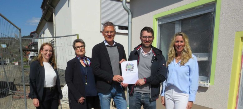 Fünf Personen stehen vor einer im Bau befindlichen Kindertagesstätte. Zwei Männer in der Mitte halten gemeinsam ein offizielles Dokument. Alle Personen blicken lächelnd in die Kamera. Im Hintergrund ist das helle Gebäude mit teilweise abgeklebten Fenstern zu sehen.
