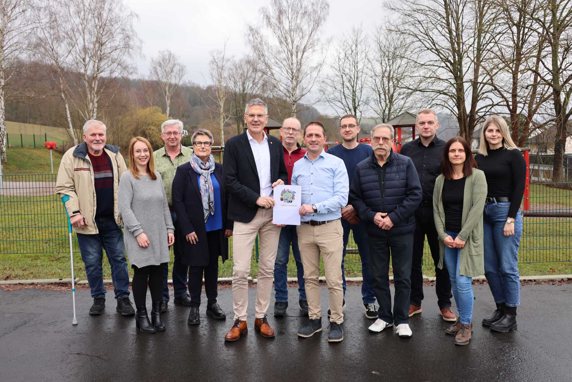 Eine größere Gruppe steht im Freien auf Asphaltboden. Zwei Männer in der Mitte halten ein Dokument mit einem bunten Motiv. Die Gruppe posiert gemeinsam für ein Foto, im Hintergrund Bäume ohne Laub.