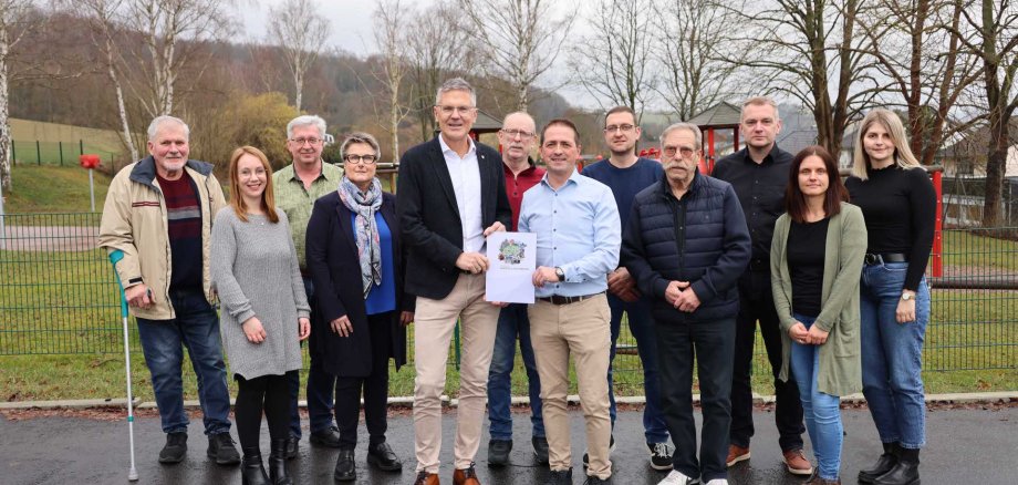 Eine größere Gruppe steht im Freien auf Asphaltboden. Zwei Männer in der Mitte halten ein Dokument mit einem bunten Motiv. Die Gruppe posiert gemeinsam für ein Foto, im Hintergrund Bäume ohne Laub.