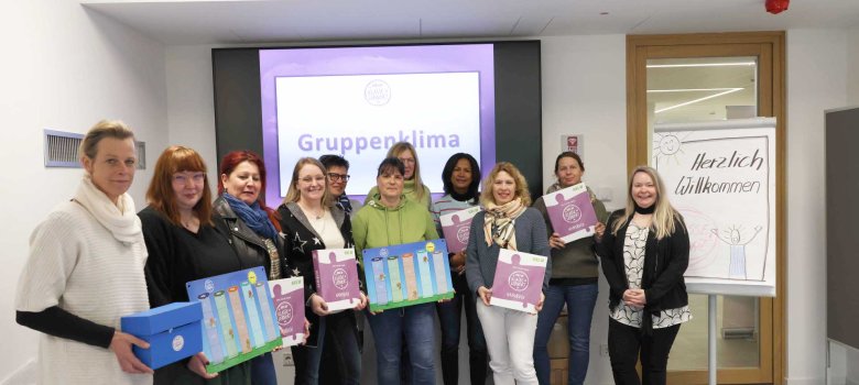 Zwölf Frauen stehen in einem Seminarraum vor einer Leinwand mit der Aufschrift „Gruppenklima“. Sie halten bunte Ordner und Materialien des Programms „Klasse Lernort“ in den Händen. Daneben steht ein Flipchart mit der Aufschrift „Herzlich Willkommen“.