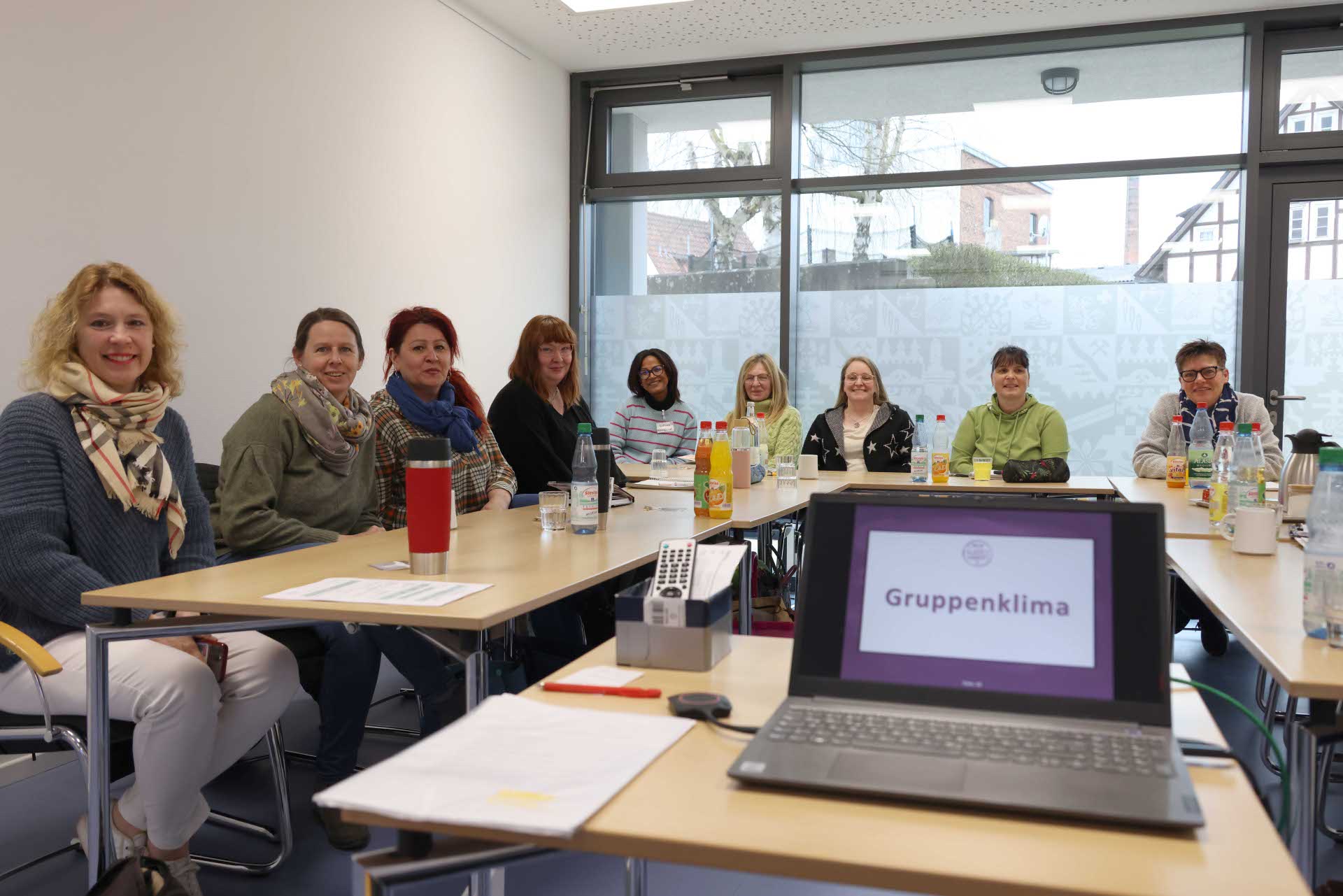 Eine Gruppe von elf Frauen sitzt an einem Konferenztisch in einem hellen Raum mit großen Fenstern. Auf dem Tisch stehen Getränke, Tassen, Unterlagen und ein Laptop, auf dessen Bildschirm „Gruppenklima“ zu lesen ist. Alle Frauen blicken freundlich in die Kamera.