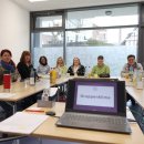 Eine Gruppe von elf Frauen sitzt an einem Konferenztisch in einem hellen Raum mit großen Fenstern. Auf dem Tisch stehen Getränke, Tassen, Unterlagen und ein Laptop, auf dessen Bildschirm „Gruppenklima“ zu lesen ist. Alle Frauen blicken freundlich in die Kamera.