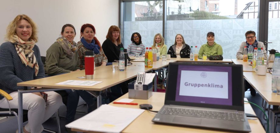 Eine Gruppe von elf Frauen sitzt an einem Konferenztisch in einem hellen Raum mit großen Fenstern. Auf dem Tisch stehen Getränke, Tassen, Unterlagen und ein Laptop, auf dessen Bildschirm „Gruppenklima“ zu lesen ist. Alle Frauen blicken freundlich in die Kamera.