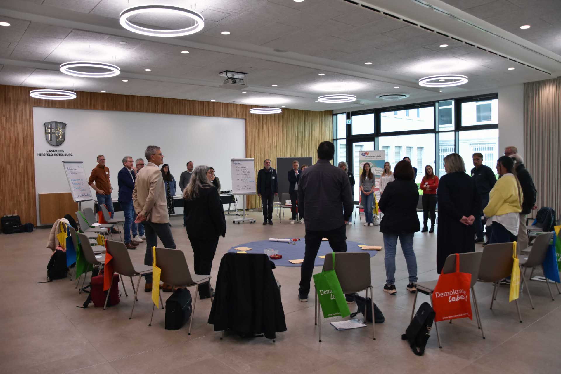 Eine Gruppe von Menschen steht im Kreis in einem modernen Konferenzraum mit großen Fenstern und hellem Licht. In der Mitte des Kreises liegen Gegenstände auf einem blauen Tuch. Um den Kreis herum stehen Stühle mit bunten Stofftaschen, auf denen „Demokratie leben!“ steht. Im Hintergrund befinden sich Flipcharts, Präsentationswände und ein Logo mit der Aufschrift „Landkreis Hersfeld-Rotenburg“. Die Atmosphäre wirkt kooperativ und partizipativ, typisch für einen Workshop oder eine Gruppenarbeit.