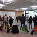 Eine Gruppe von Menschen steht im Kreis in einem modernen Konferenzraum mit großen Fenstern und hellem Licht. In der Mitte des Kreises liegen Gegenstände auf einem blauen Tuch. Um den Kreis herum stehen Stühle mit bunten Stofftaschen, auf denen „Demokratie leben!“ steht. Im Hintergrund befinden sich Flipcharts, Präsentationswände und ein Logo mit der Aufschrift „Landkreis Hersfeld-Rotenburg“. Die Atmosphäre wirkt kooperativ und partizipativ, typisch für einen Workshop oder eine Gruppenarbeit.