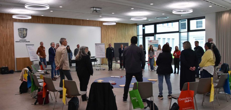 Eine Gruppe von Menschen steht im Kreis in einem modernen Konferenzraum mit großen Fenstern und hellem Licht. In der Mitte des Kreises liegen Gegenstände auf einem blauen Tuch. Um den Kreis herum stehen Stühle mit bunten Stofftaschen, auf denen „Demokratie leben!“ steht. Im Hintergrund befinden sich Flipcharts, Präsentationswände und ein Logo mit der Aufschrift „Landkreis Hersfeld-Rotenburg“. Die Atmosphäre wirkt kooperativ und partizipativ, typisch für einen Workshop oder eine Gruppenarbeit.