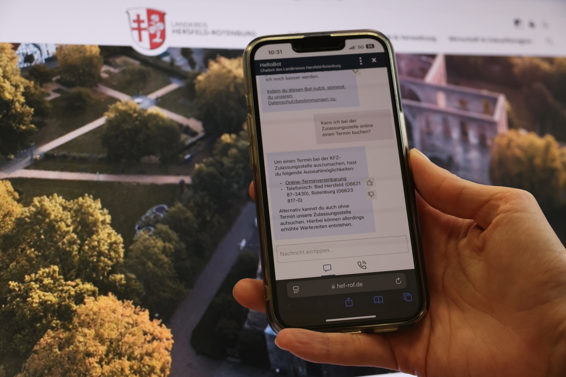 Chatbot HeRoBot Smartphone zeigt den Chatbot „HeRoBot“ des Landkreises Hersfeld-Rotenburg bei der Beantwortung einer Bürgerfrage zur Terminvereinbarung bei der KFZ-Zulassungsstelle. Im Hintergrund: Startseite der Landkreis-Website mit dem Logo und einem Luftbild des Bad Hersfelder Stiftbezirks.