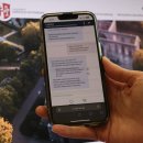 Smartphone zeigt den Chatbot „HeRoBot“ des Landkreises Hersfeld-Rotenburg bei der Beantwortung einer Bürgerfrage zur Terminvereinbarung bei der KFZ-Zulassungsstelle. Im Hintergrund: Startseite der Landkreis-Website mit dem Logo und einem Luftbild des Bad Hersfelder Stiftbezirks.
