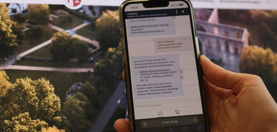 Smartphone zeigt den Chatbot „HeRoBot“ des Landkreises Hersfeld-Rotenburg bei der Beantwortung einer Bürgerfrage zur Terminvereinbarung bei der KFZ-Zulassungsstelle. Im Hintergrund: Startseite der Landkreis-Website mit dem Logo und einem Luftbild des Bad Hersfelder Stiftbezirks.