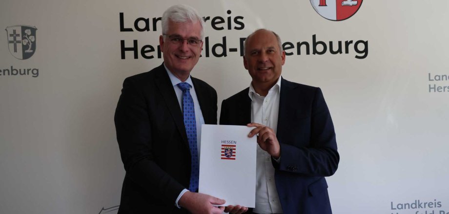 Zwei Männer stehen nebeneinander vor einer Wand mit dem Logo des Landkreises Hersfeld-Rotenburg. Sie halten gemeinsam eine Urkunde in der Hand. Links steht Landrat Torsten Warnecke im Anzug, rechts Hessens Innenminister Roman Poseck ebenfalls im Anzug. Die beiden blicken in die Kamera und lächeln.