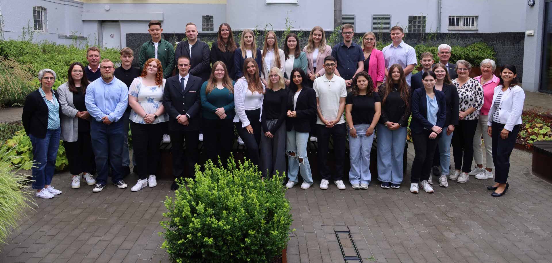 Ein Gruppenfoto mit etwa 35 Personen, aufgenommen im Innenhof eines Bürogebäudes. Die Gruppe besteht aus einer Mischung aus jungen Erwachsenen und älteren Personen – vermutlich neue Auszubildende und Mitarbeitende eines Unternehmens. Die Personen stehen in zwei Reihen: Die hintere Reihe steht auf einer leicht erhöhten Plattform, die vordere Reihe steht auf dem Boden. Im Hintergrund sieht man die Fassade des Gebäudes mit mehreren Fenstern und Klimaanlagen. Die Personen sind überwiegend leger bis halbformell gekleidet, einige tragen Blazer, Hemden oder Businesskleidung. Es herrscht eine freundliche und professionelle Stimmung.