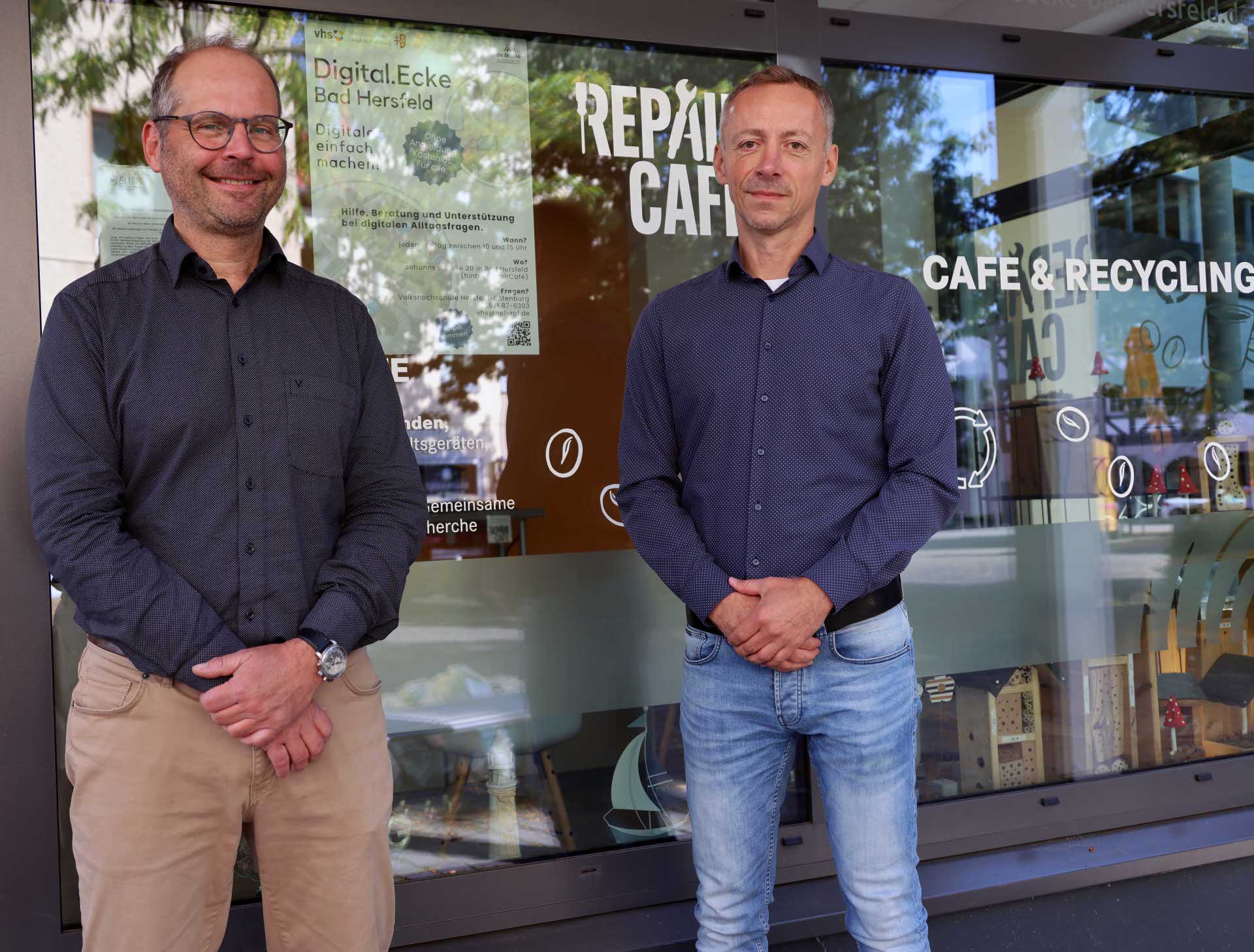 Zwei Männer stehen nebeneinander vor der Glasfassade eines Cafés mit der Aufschrift „Repair Café – Café & Recycling“. Im Fenster hängt ein Plakat zur „Digital.Ecke Bad Hersfeld“.