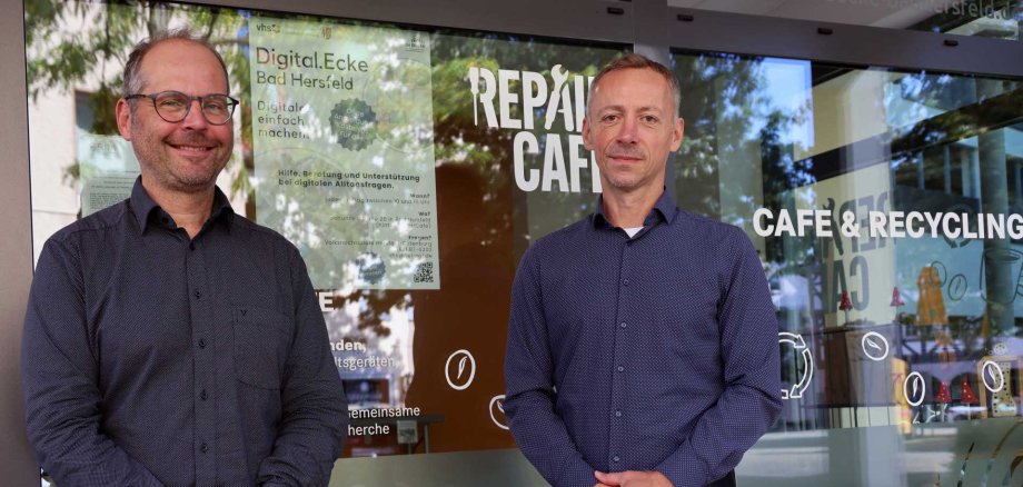 Zwei Männer stehen nebeneinander vor der Glasfassade eines Cafés mit der Aufschrift „Repair Café – Café & Recycling“. Im Fenster hängt ein Plakat zur „Digital.Ecke Bad Hersfeld“.