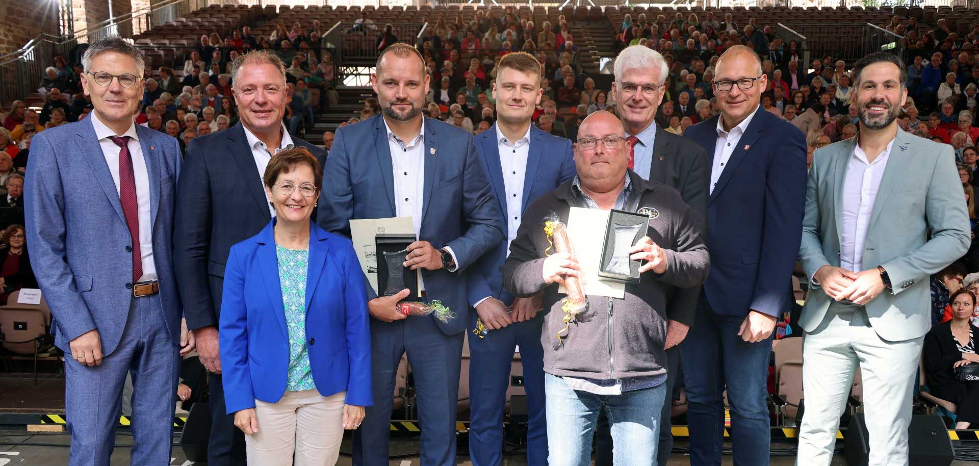 Eine Gruppe von neun Personen posiert auf einer Bühne vor einem großen Publikum. Zwei Männer in der Mitte halten Urkunden und kleine Geschenke in den Händen. Die Personen tragen überwiegend Anzüge, eine Frau in der vorderen Reihe ist in heller Hose, türkisfarbenem Oberteil und blauem Blazer gekleidet. Im Hintergrund sitzt ein dicht besetztes Publikum in einem überdachten Veranstaltungsort.