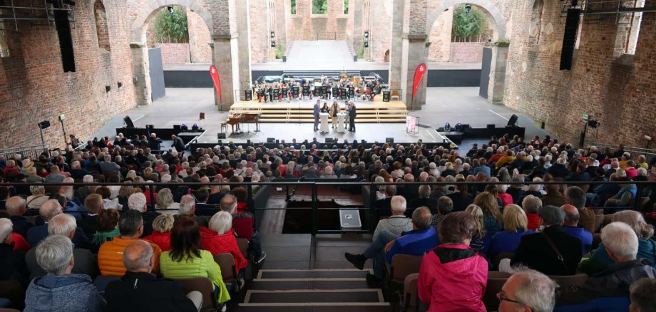 Im Vordergrund sind viele Zuschauer zu sehen. Im Hintergrund gibt es eine Bühne in einer historischen Kulisse,
