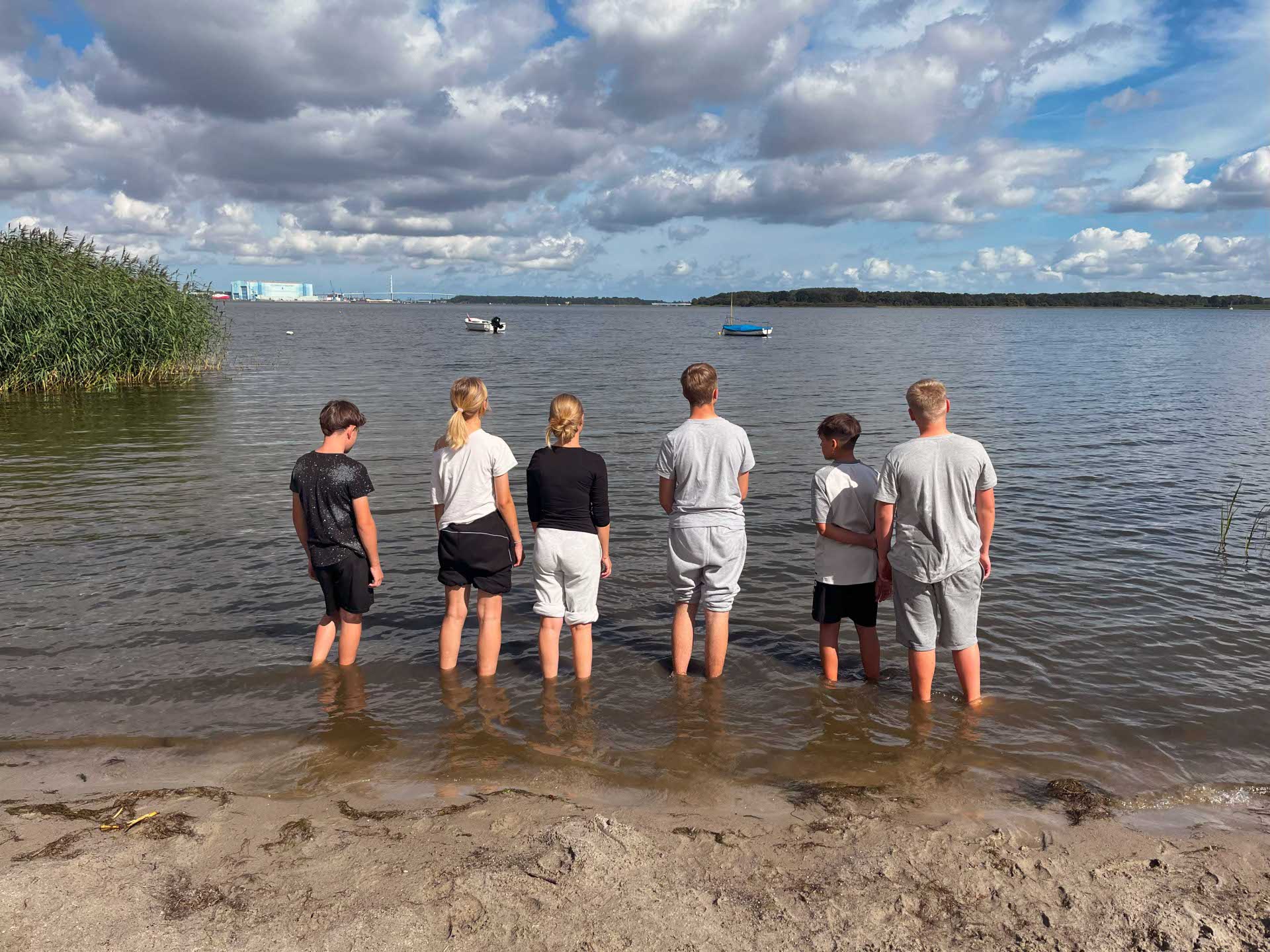 Sechs Kinder stehen barfuß am Ufer eines Sees und blicken auf das Wasser hinaus. Am Horizont sind Boote, eine grüne Insel und Industriegebäude zu sehen. Der Himmel ist von vielen Wolken bedeckt, durch die teilweise die Sonne scheint.