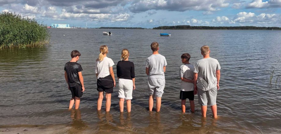 Sechs Kinder stehen barfuß am Ufer eines Sees und blicken auf das Wasser hinaus. Am Horizont sind Boote, eine grüne Insel und Industriegebäude zu sehen. Der Himmel ist von vielen Wolken bedeckt, durch die teilweise die Sonne scheint.