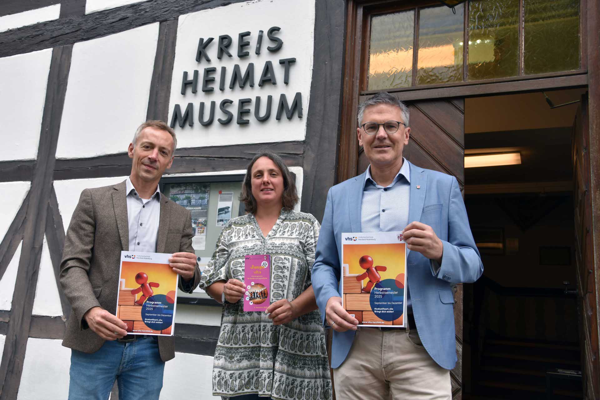 Drei Personen stehen vor dem Eingang eines Fachwerkgebäudes mit dem Schild „Kreis Heimat Museum“. Sie halten Broschüren in den Händen. Die beiden Männer links und rechts tragen Anzüge und halten jeweils ein Programmheft der Volkshochschule (vhs) für das Herbstsemester 2025. Die Frau in der Mitte trägt ein gemustertes Kleid und hält ein Heft mit dem Titel „Junge vhs“. Im Hintergrund ist der geöffnete Eingang des Museums zu sehen.