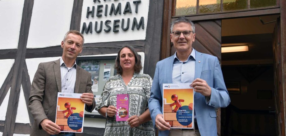 Drei Personen stehen vor dem Eingang eines Fachwerkgebäudes mit dem Schild „Kreis Heimat Museum“. Sie halten Broschüren in den Händen. Die beiden Männer links und rechts tragen Anzüge und halten jeweils ein Programmheft der Volkshochschule (vhs) für das Herbstsemester 2025. Die Frau in der Mitte trägt ein gemustertes Kleid und hält ein Heft mit dem Titel „Junge vhs“. Im Hintergrund ist der geöffnete Eingang des Museums zu sehen.