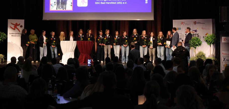 Einige Frauen stehen auf einer Bühne. Sie halten Blumen in der Hand. Auf einer Leinwand werden drei Mannschaften gezeigt. Ein Moderator hält ein Mikrofon in der Hand.