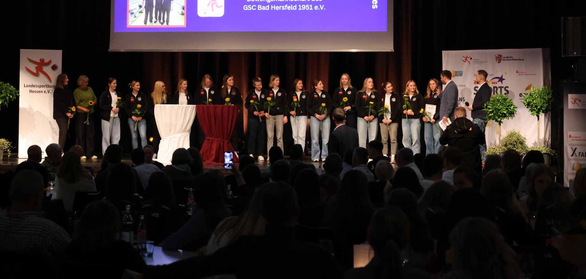 Einige Frauen stehen auf einer Bühne. Sie halten Blumen in der Hand. Auf einer Leinwand werden drei Mannschaften gezeigt. Ein Moderator hält ein Mikrofon in der Hand.