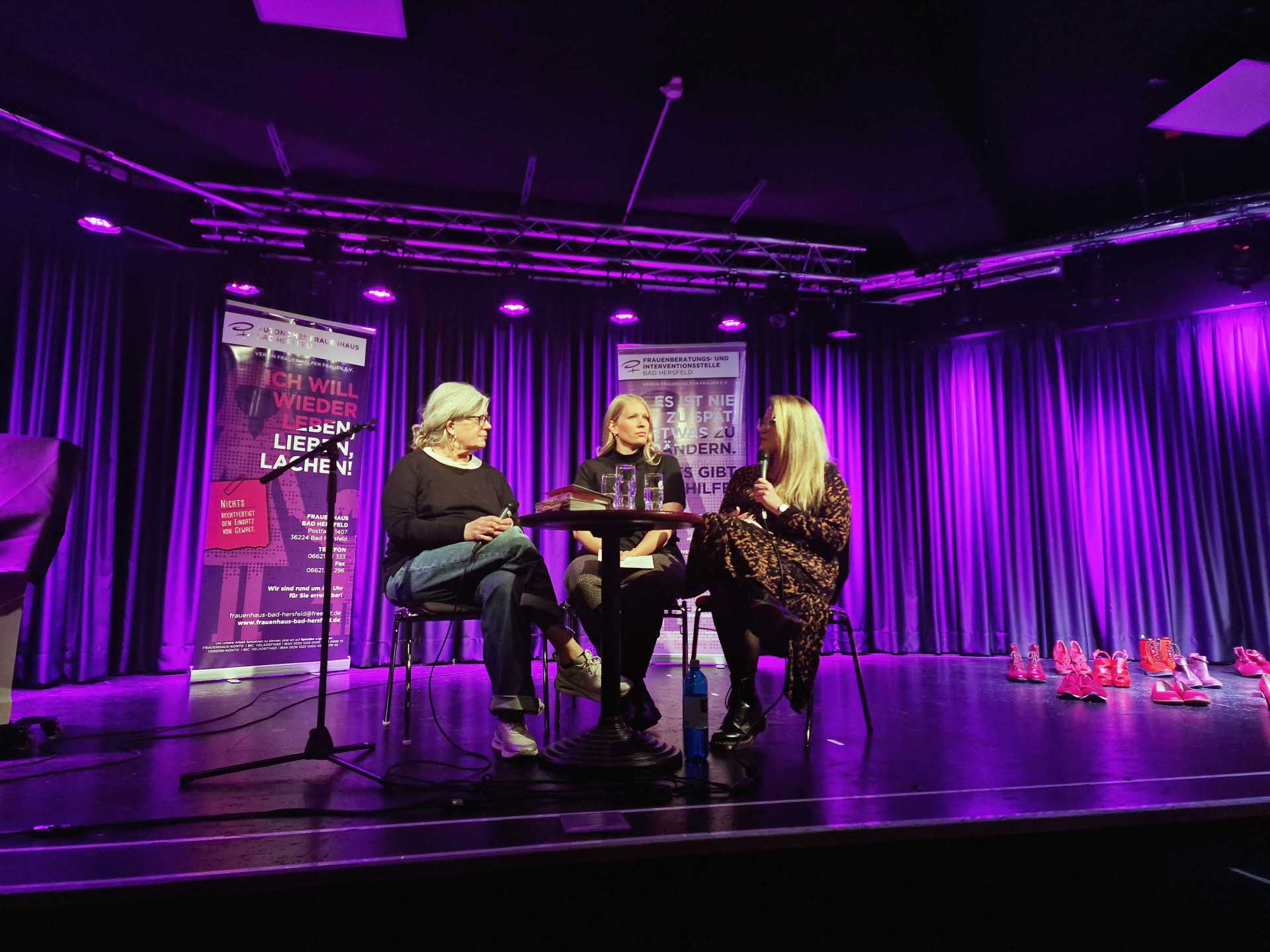Drei Frauen sitzen auf einer Bühne an einem kleinen runden Tisch und führen ein Gespräch. Hinter ihnen hängen violett beleuchtete Vorhänge und zwei Infobanner mit Text. Rechts am Bühnenrand stehen viele pinke und rote Frauenschuhe in Reihen. Die Szene wirkt wie eine öffentliche Lesung oder Podiumsdiskussion in einem Veranstaltungsraum.