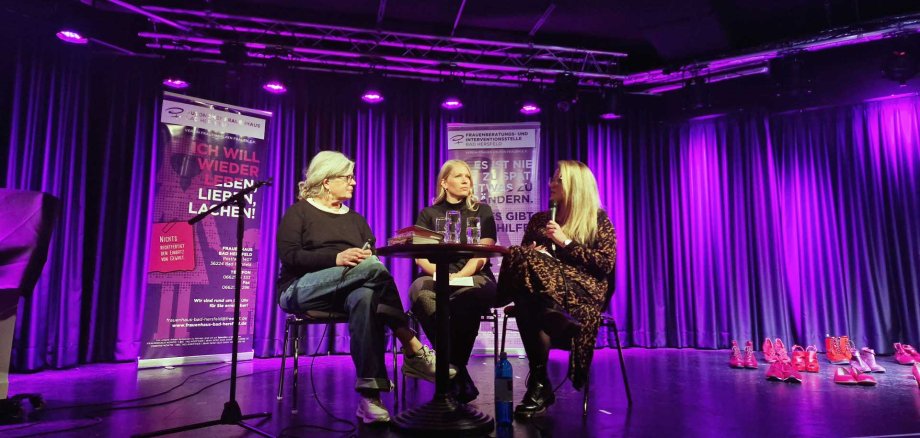 Drei Frauen sitzen auf einer Bühne an einem kleinen runden Tisch und führen ein Gespräch. Hinter ihnen hängen violett beleuchtete Vorhänge und zwei Infobanner mit Text. Rechts am Bühnenrand stehen viele pinke und rote Frauenschuhe in Reihen. Die Szene wirkt wie eine öffentliche Lesung oder Podiumsdiskussion in einem Veranstaltungsraum.