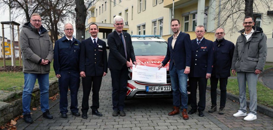 Eine Gruppe von acht Männern steht im Freien vor einem rot-weißen Einsatzfahrzeug der Feuerwehr. Zwei Männer in der Mitte halten gemeinsam einen großen symbolischen Scheck. Einige der Männer tragen Feuerwehr- oder Dienstuniformen, andere tragen zivile Kleidung. Im Hintergrund sind ein Gebäude und winterliche Bäume zu sehen.