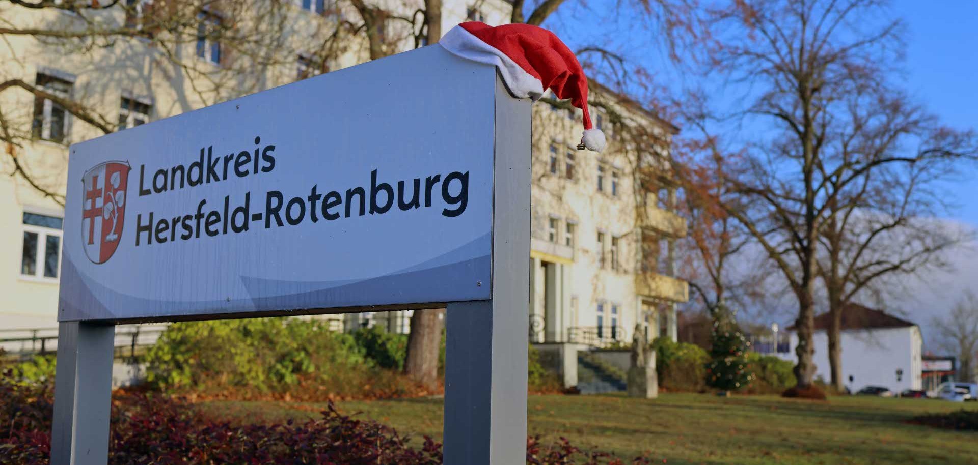 Straßenschild mit der Aufschrift Landkreis Hersfeld-Rotenburg und Wappen, auf dem eine Weihnachtsmütze liegt. Im Hintergrund stehen ein helles Verwaltungsgebäude, kahle Bäume und ein Weihnachtsbaum auf einer grünen Rasenfläche unter blauem Himmel.