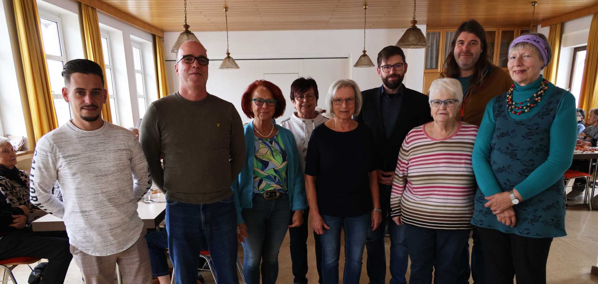 Eine Gruppe von zehn Personen unterschiedlichen Alters steht in einem Gemeinschaftsraum in zwei Reihen nebeneinander. Sie blicken lächelnd in die Kamera. Im Hintergrund sitzen weitere Menschen an Tischen, und der Raum ist hell mit großen Fenstern und gelben Vorhängen.
