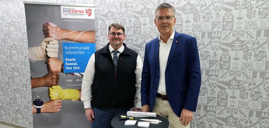 Zwei Männer stehen in einem Innenraum vor einer grauen Wand mit grafischem Muster. Links befindet sich ein Roll-Up-Banner des kommunalen Jobcenters mit dem Slogan „Stark. Sozial. Vor Ort.“ und mehreren aneinanderstoßenden Fäusten unterschiedlicher Hände. Die beiden Männer stehen neben einem kleinen runden Stehtisch mit Werbematerialien des Jobcenters und blicken lächelnd in die Kamera.