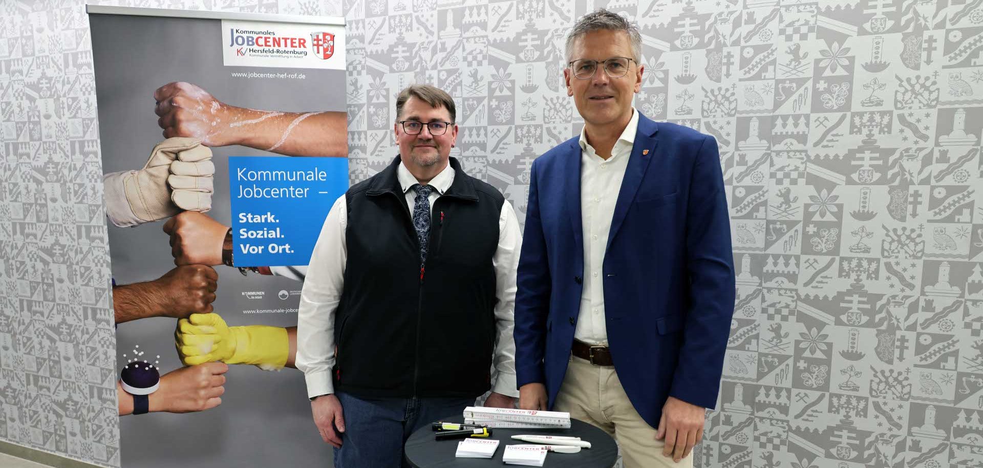 Zwei Männer stehen in einem Innenraum vor einer grauen Wand mit grafischem Muster. Links befindet sich ein Roll-Up-Banner des kommunalen Jobcenters mit dem Slogan „Stark. Sozial. Vor Ort.“ und mehreren aneinanderstoßenden Fäusten unterschiedlicher Hände. Die beiden Männer stehen neben einem kleinen runden Stehtisch mit Werbematerialien des Jobcenters und blicken lächelnd in die Kamera.