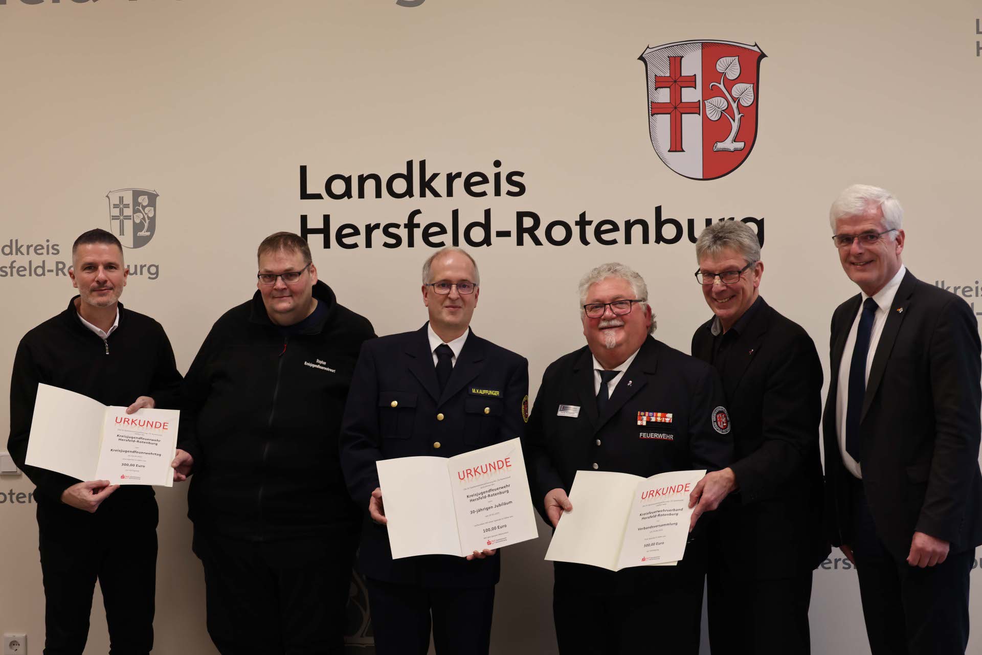 Mehrere Männer stehen gemeinsam vor einer Wand mit dem Logo des Landkreises Hersfeld-Rotenburg. Drei Männer halten geöffnete Urkunden in den Händen.