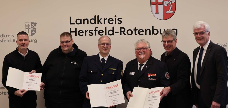 Mehrere Männer stehen gemeinsam vor einer Wand mit dem Logo des Landkreises Hersfeld-Rotenburg. Drei Männer halten geöffnete Urkunden in den Händen.