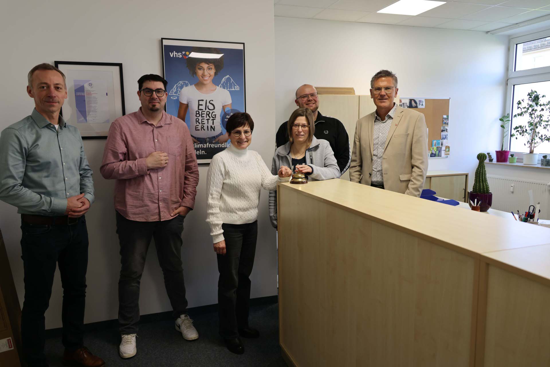 Mehrere Personen stehen hinter und vor einem hellen Empfangstresen in den neuen Räumen der Volkshochschule, im Hintergrund sind Büromöbel und Infomaterial zu sehen.