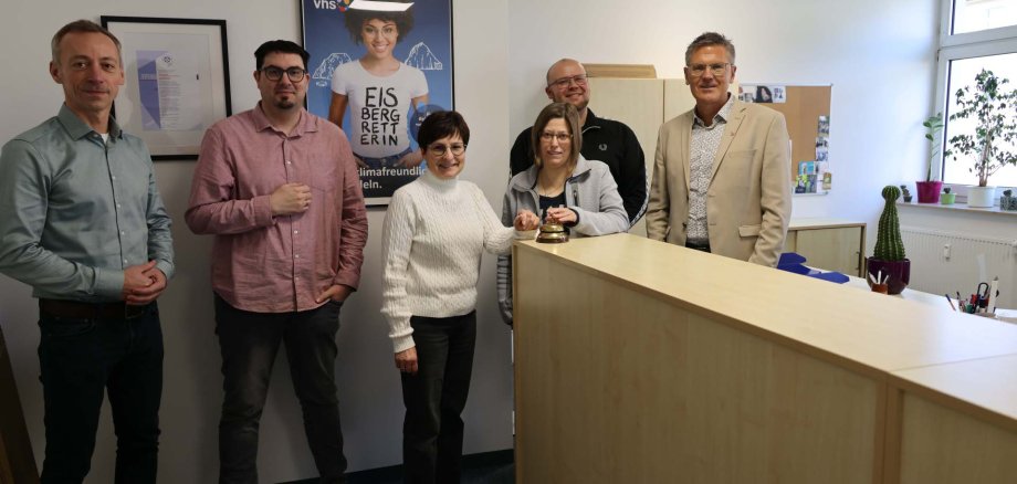 Mehrere Personen stehen hinter und vor einem hellen Empfangstresen in den neuen Räumen der Volkshochschule, im Hintergrund sind Büromöbel und Infomaterial zu sehen.