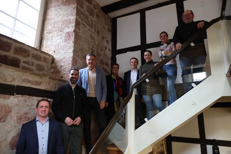 Eine Gruppe von acht Personen steht auf einer Treppe in einem historischen Fachwerkgebäude. Die Personen sind in verschiedenen Positionen verteilt, einige stehen auf der Treppe, andere daneben. Die Gruppe besteht aus Vertretern des Landkreises, der Kommunen sowie der Drogenhilfe Nordhessen e.V., die sich für das SoS-Projekt engagieren.