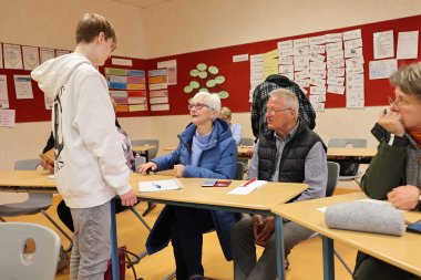 Ein Klassenzimmer mit mehreren Seniorinnen und Senioren, die an Schultischen sitzen. Ein Jugendlicher mit weißem Hoodie steht vor einer älteren Frau mit Brille und blauer Jacke und erklärt ihr etwas. Weitere Teilnehmer hören zu oder notieren sich Informationen.