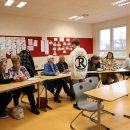 Mehrere Gruppen aus Seniorinnen, Senioren und Jugendlichen sitzen in einem Klassenzimmer. Sie diskutieren und arbeiten gemeinsam an digitalen Themen. Im Hintergrund sind Lernmaterialien an einer roten Pinnwand und große Fenster, die Tageslicht hereinlassen.