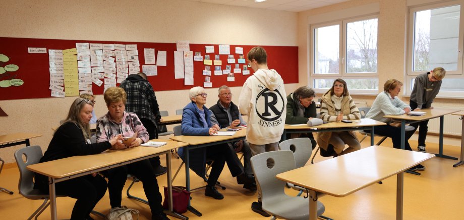 Mehrere Gruppen aus Seniorinnen, Senioren und Jugendlichen sitzen in einem Klassenzimmer. Sie diskutieren und arbeiten gemeinsam an digitalen Themen. Im Hintergrund sind Lernmaterialien an einer roten Pinnwand und große Fenster, die Tageslicht hereinlassen.