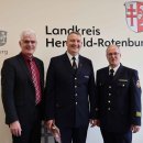 Drei Männer stehen vor einer Tapete mit der Aufschrift "Landkreis Hersfeld-Rotenburg". Zwei von ihnen tragen eine Feuerwehruniform. Der Mann in der Mitte hält eine Urkunde und eine Stracke.