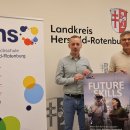 Fabian Göbel, Leiter der Volkshochschule, und Dirk Noll, Erster Kreisbeigeordneter, stehen vor einem Banner der Volkshochschule (VHS) Hersfeld-Rotenburg und einem Schild mit dem Wappen und Schriftzug des Landkreises Hersfeld-Rotenburg. Fabian Göbel hält ein Poster mit der Aufschrift „Future Skills“ und einer Abbildung von Personen mit VR-Brille. Dirk Noll präsentiert das Programmheft der VHS für das Frühjahrsemester 2025. Das Banner trägt den Slogan „Hätte... Sollte... Könnte... Würde... Machen!“ und zeigt bunte Kreise als Gestaltungselemente.