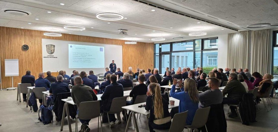 Ein großer Konferenzraum mit mehreren Reihen von Teilnehmern, die an Tischen sitzen und auf einen Vortragenden vorne blicken. An der Wand wird eine Präsentation mit dem Titel „Ausgangslage“ projiziert, die verschiedene Themen anspricht. Der Raum hat moderne Einrichtung mit Holzverkleidung an der Vorderwand, einer Uhr und dem Wappen des Landkreises Hersfeld-Rotenburg.